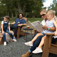 A family of four enjoys a relaxing patio gathering outdoors. The mother reads to a young girl on her lap in a Ledge Lounger Legacy Adirondack chair, while another girl sits on a step. The father, seated nearby with a drink, is surrounded by stacks of firewood and trees in the background.