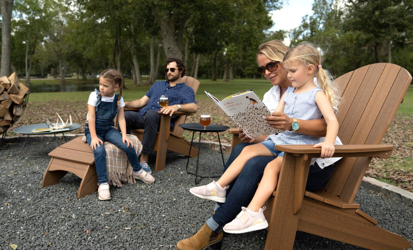 A family of four enjoys a relaxing patio gathering outdoors. The mother reads to a young girl on her lap in a Ledge Lounger Legacy Adirondack chair, while another girl sits on a step. The father, seated nearby with a drink, is surrounded by stacks of firewood and trees in the background.