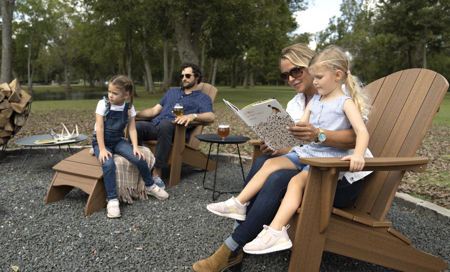 A family of four enjoys a relaxing patio gathering outdoors. The mother reads to a young girl on her lap in a Ledge Lounger Legacy Adirondack chair, while another girl sits on a step. The father, seated nearby with a drink, is surrounded by stacks of firewood and trees in the background.