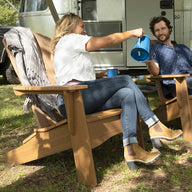 A woman and a man unwind in Ledge Loungers Legacy Adirondack chairs outside an Airstream trailer. Shes in a white top and jeans, pouring from a blue thermos, while he wears a blue shirt and jeans, smiling warmly. Nearby, under towering trees on the grass, sits a parked black truck.