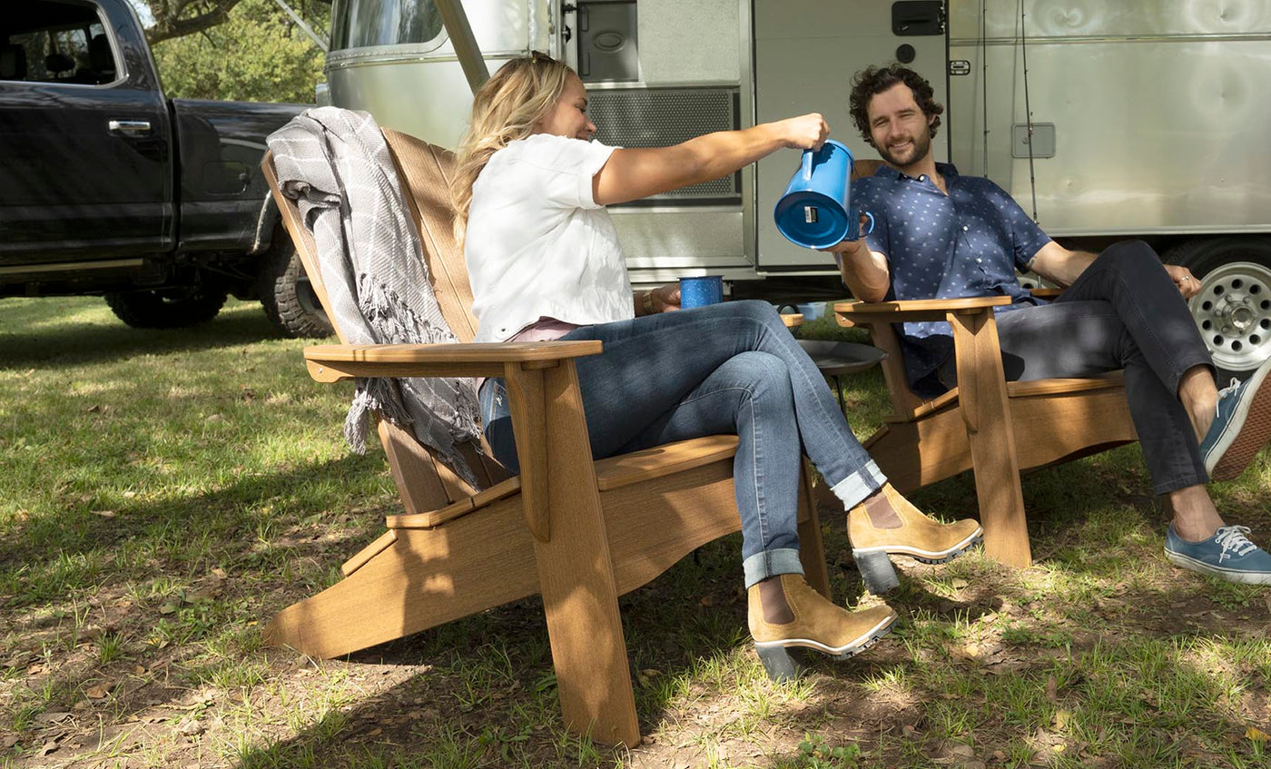 A woman and a man unwind in Ledge Loungers Legacy Adirondack chairs outside an Airstream trailer. Shes in a white top and jeans, pouring from a blue thermos, while he wears a blue shirt and jeans, smiling warmly. Nearby, under towering trees on the grass, sits a parked black truck.