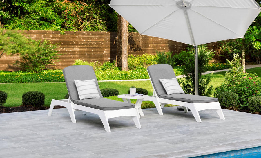 Two gray lounge chairs with striped outdoor fabric pillows sit beside a small round table under a large white umbrella on a tiled poolside patio, surrounded by a wooden fence, greenery, and trees—ideal for relaxation with Ledge Loungers customizable Legacy Chaise Cushion.