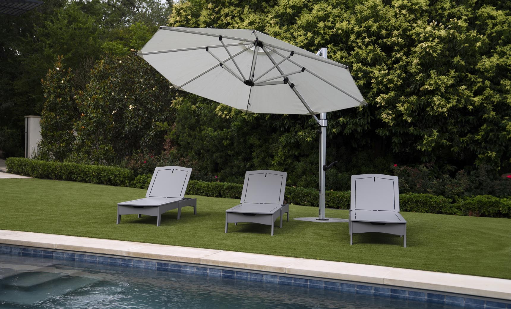 Three Ledge Lounger Legacy Chaise lounge chairs are set on green grass under a large white umbrella, adjustable to various reclining positions and facing the pool. Dense greenery with tall bushes and trees provides a serene backdrop to this tranquil outdoor scene.