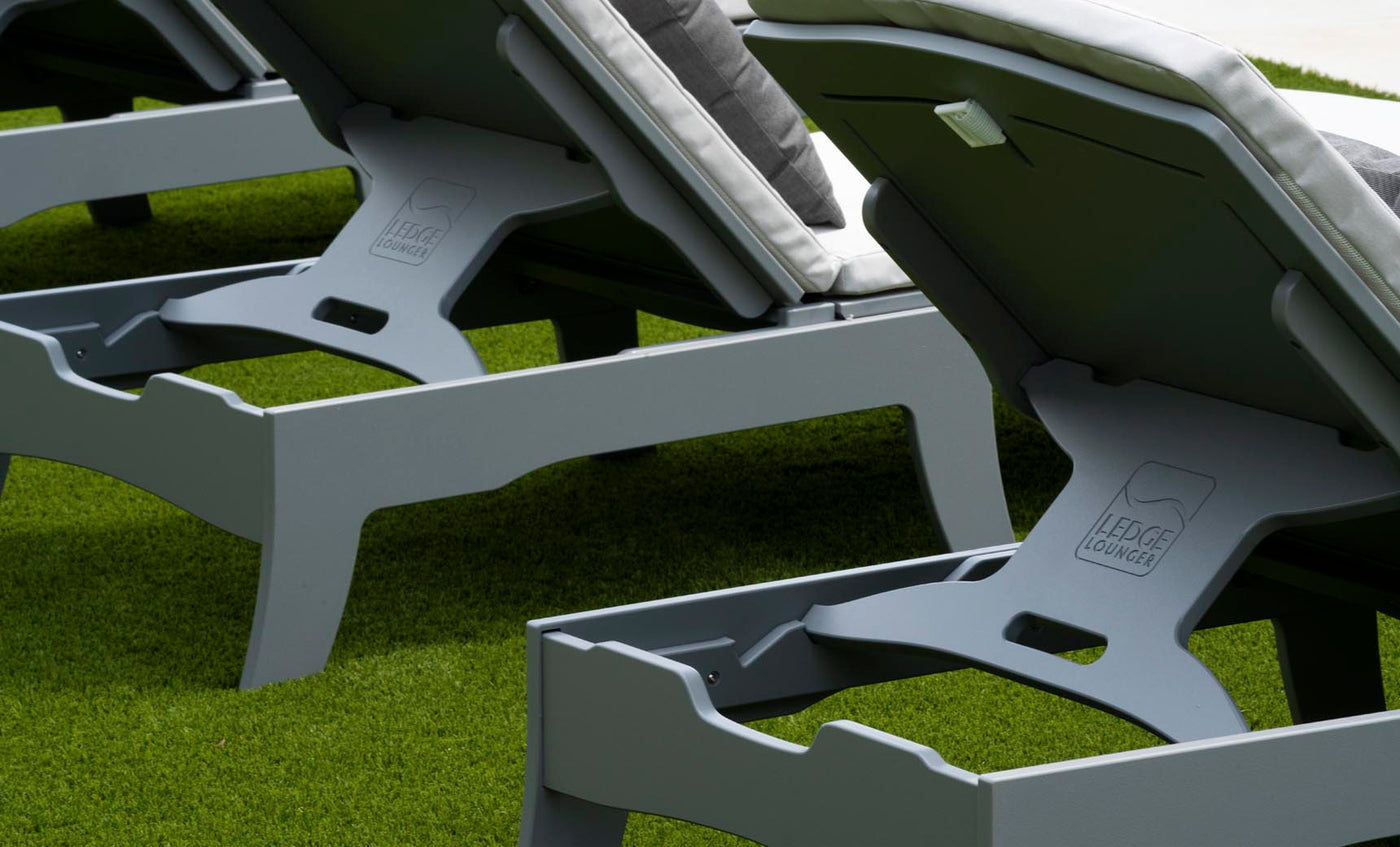 Close-up of grey Legacy Chaise lounge chairs from Ledge Lounger, lined up on green artificial turf. Featuring white cushions and multiple reclining positions, LEDGE LOUNGER is visible on the bases in this inviting outdoor space.