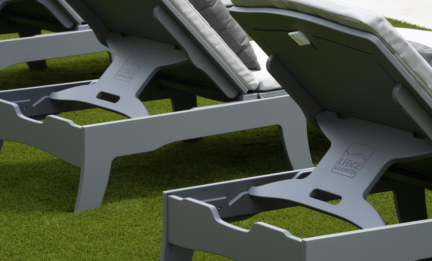 Close-up of grey Legacy Chaise lounge chairs from Ledge Lounger, lined up on green artificial turf. Featuring white cushions and multiple reclining positions, LEDGE LOUNGER is visible on the bases in this inviting outdoor space.