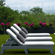 Three white Ledge Lounger Legacy Chaise lounge chairs with dark cushions are lined up on the grass in this serene outdoor setting. Vibrant purple flowers bloom in planters behind them, with a city skyline visible under an overcast sky.