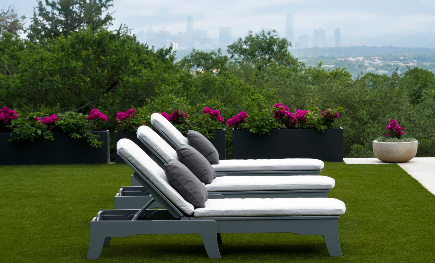 Three white Ledge Lounger Legacy Chaise lounge chairs with dark cushions are lined up on the grass in this serene outdoor setting. Vibrant purple flowers bloom in planters behind them, with a city skyline visible under an overcast sky.
