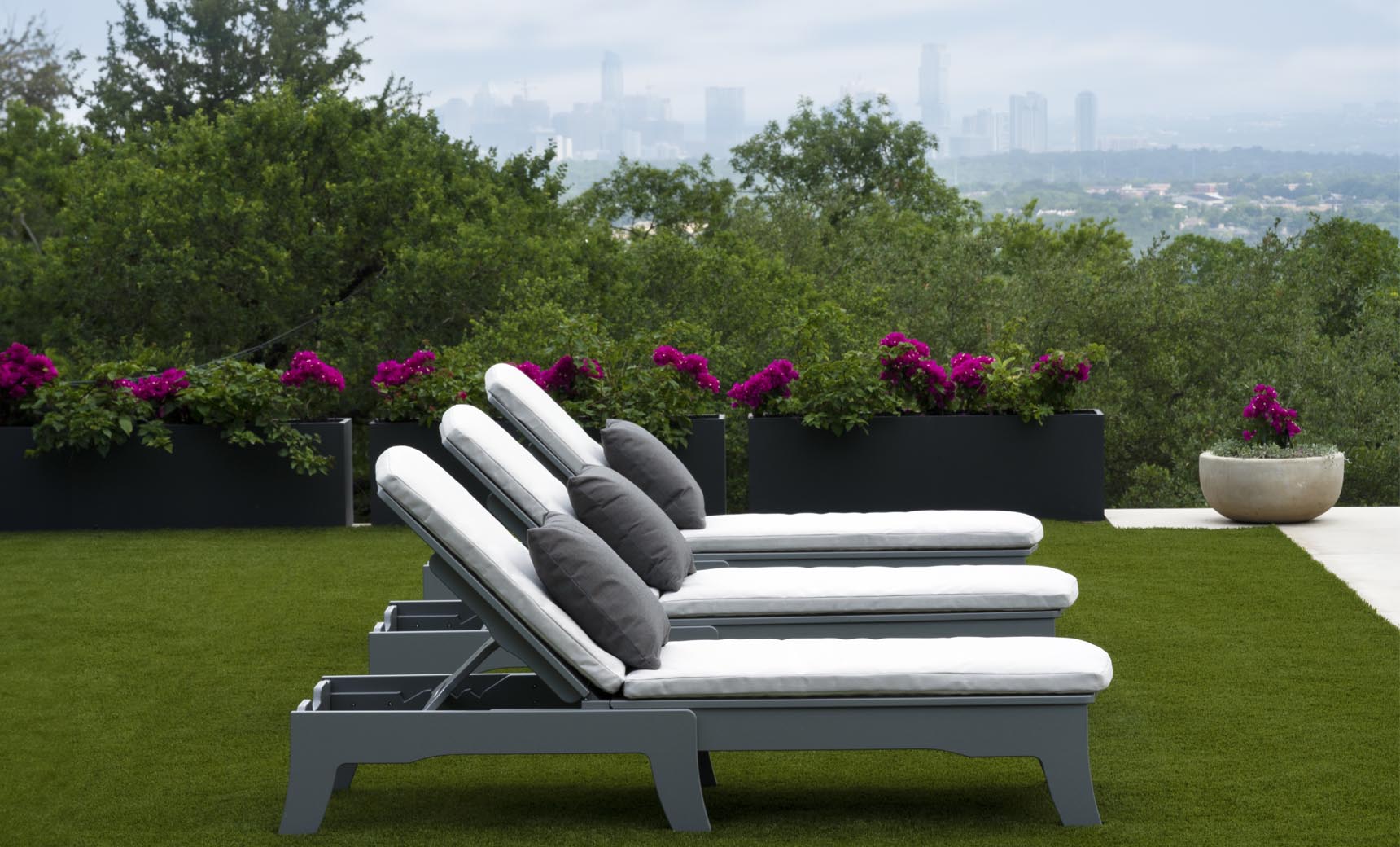 Three white Ledge Lounger Legacy Chaise lounge chairs with dark cushions are lined up on the grass in this serene outdoor setting. Vibrant purple flowers bloom in planters behind them, with a city skyline visible under an overcast sky.