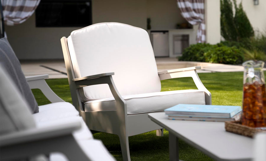 A white Legacy Club Chair from Ledge Lounger sits cushioned on the green lawn, creating a cozy outdoor space. Behind it, an outdoor kitchen with striped curtains adds charm, while a nearby table holds a book and a jug of iced tea, inviting relaxation and leisure in this customizable design.