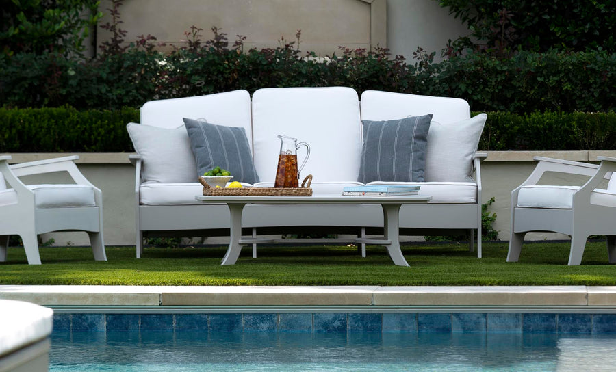 The Ledge Lounger Legacy Sofa, a traditional deep seat design, features durable white outdoor fabrics with gray cushions by the pool. Nearby, a coffee table holds refreshments and magazines on a wicker tray, while lush greenery and bushes frame the serene scene.