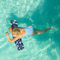 A blonde person in a blue swimsuit relaxes on the Luxurious Laze-y Days Float Bundle by Ledge Lounger in clear turquoise water, with sunlight dancing across the surface creating a serene tropical vibe.