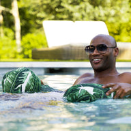 A person in sunglasses smiles while relaxing in a pool, enjoying a green tropical-patterned Laze Hammock made of marine-grade fabrics by Ledge Lounger. Blurred greenery and a sunlit lounge chair enhance the vibe of outdoor adventures.