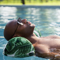 A man in sunglasses relaxes in a Ledge Lounger Laze Hammock float made with marine-grade fabrics, drifting in a calm pool. His eyes are closed and head tilted back as the water blends seamlessly with the blurred trees and grass around him.