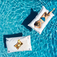 A person in a blue swimsuit lounges on a white inflatable float in a clear pool. Beside them, the Laze Pillow by Ledge Lounger supports a pizza and woven bag. Sunlight shimmers on the water, creating gentle ripples.