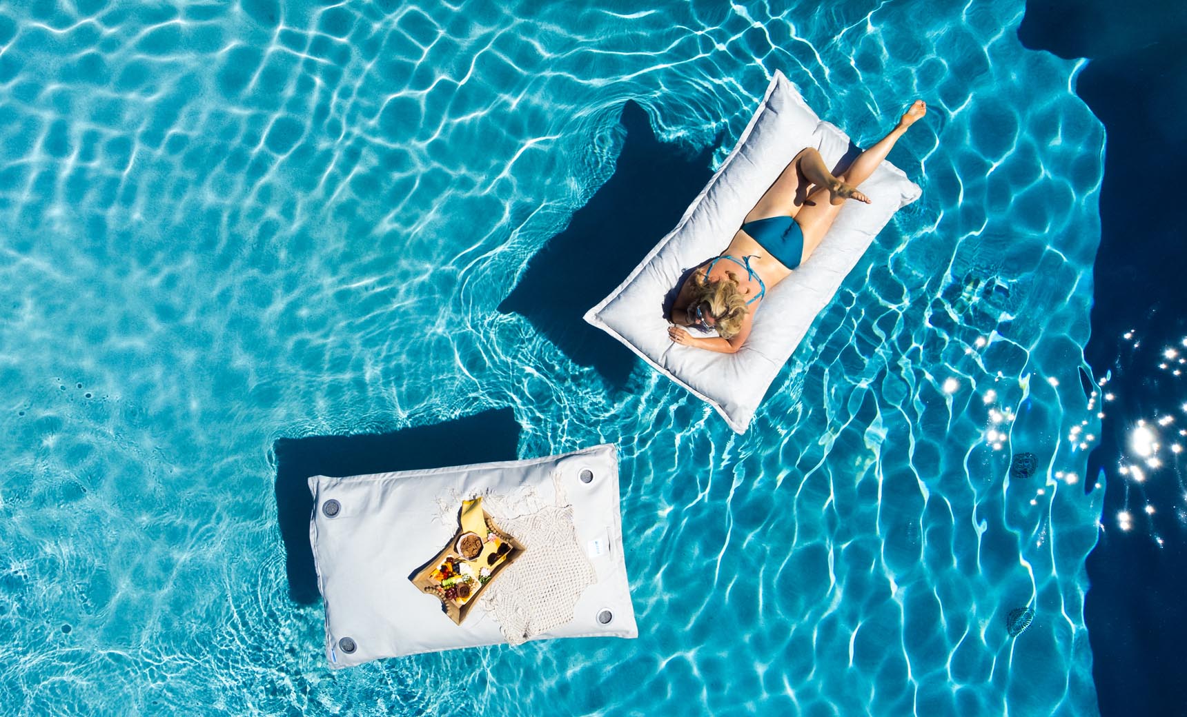 A person in a blue swimsuit lounges on a white inflatable float in a clear pool. Beside them, the Laze Pillow by Ledge Lounger supports a pizza and woven bag. Sunlight shimmers on the water, creating gentle ripples.