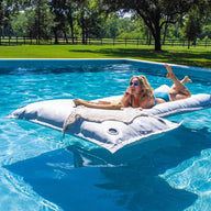 Lounging outdoors on Ledge Lounger’s Laze Pillow, a person in sunglasses and swimsuit relaxes stomach-down on the large white inflatable in a blue pool. The green grass and trees under the clear sky perfectly complement this floating cushion experience.