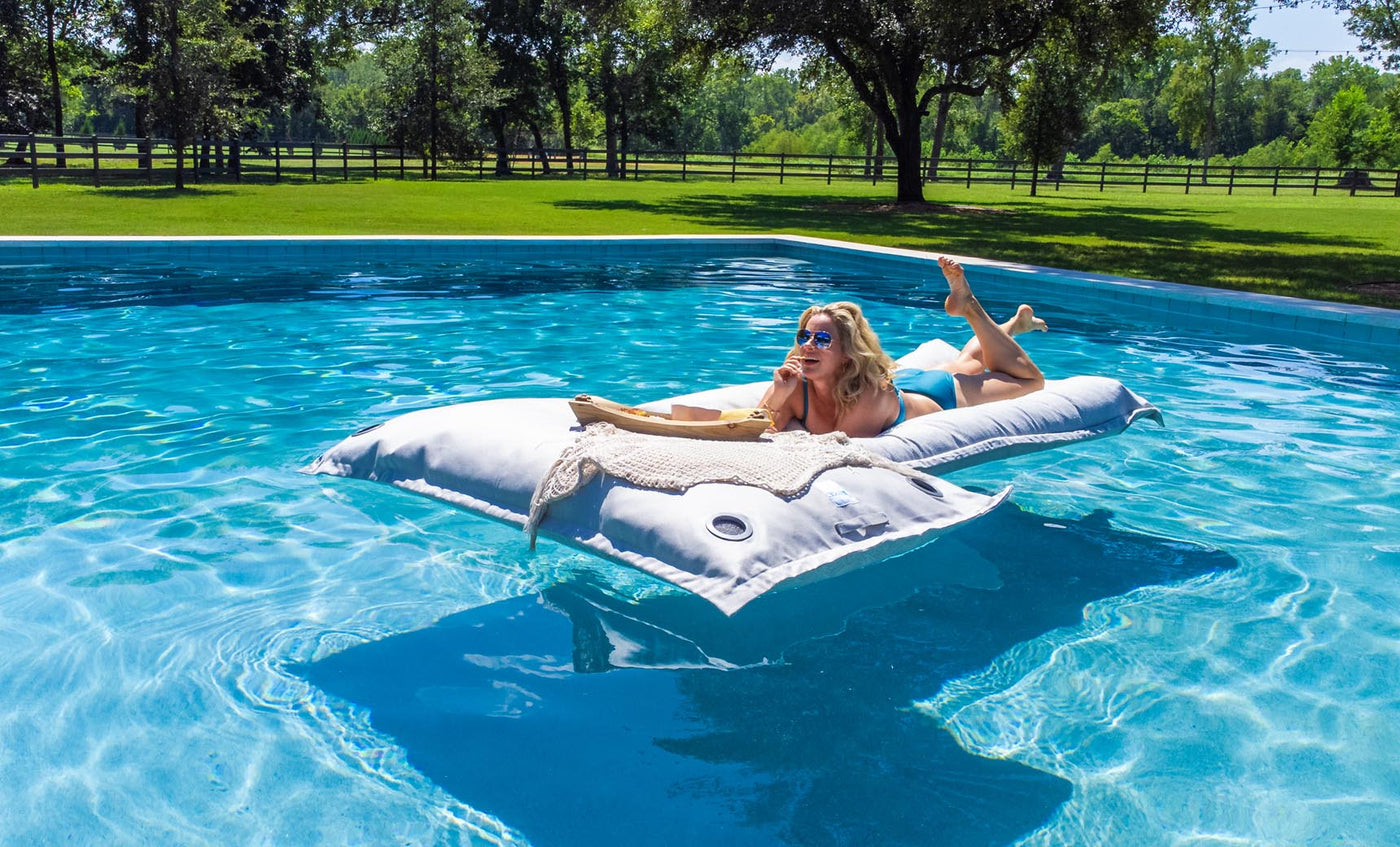 Lounging outdoors on Ledge Lounger’s Laze Pillow, a person in sunglasses and swimsuit relaxes stomach-down on the large white inflatable in a blue pool. The green grass and trees under the clear sky perfectly complement this floating cushion experience.