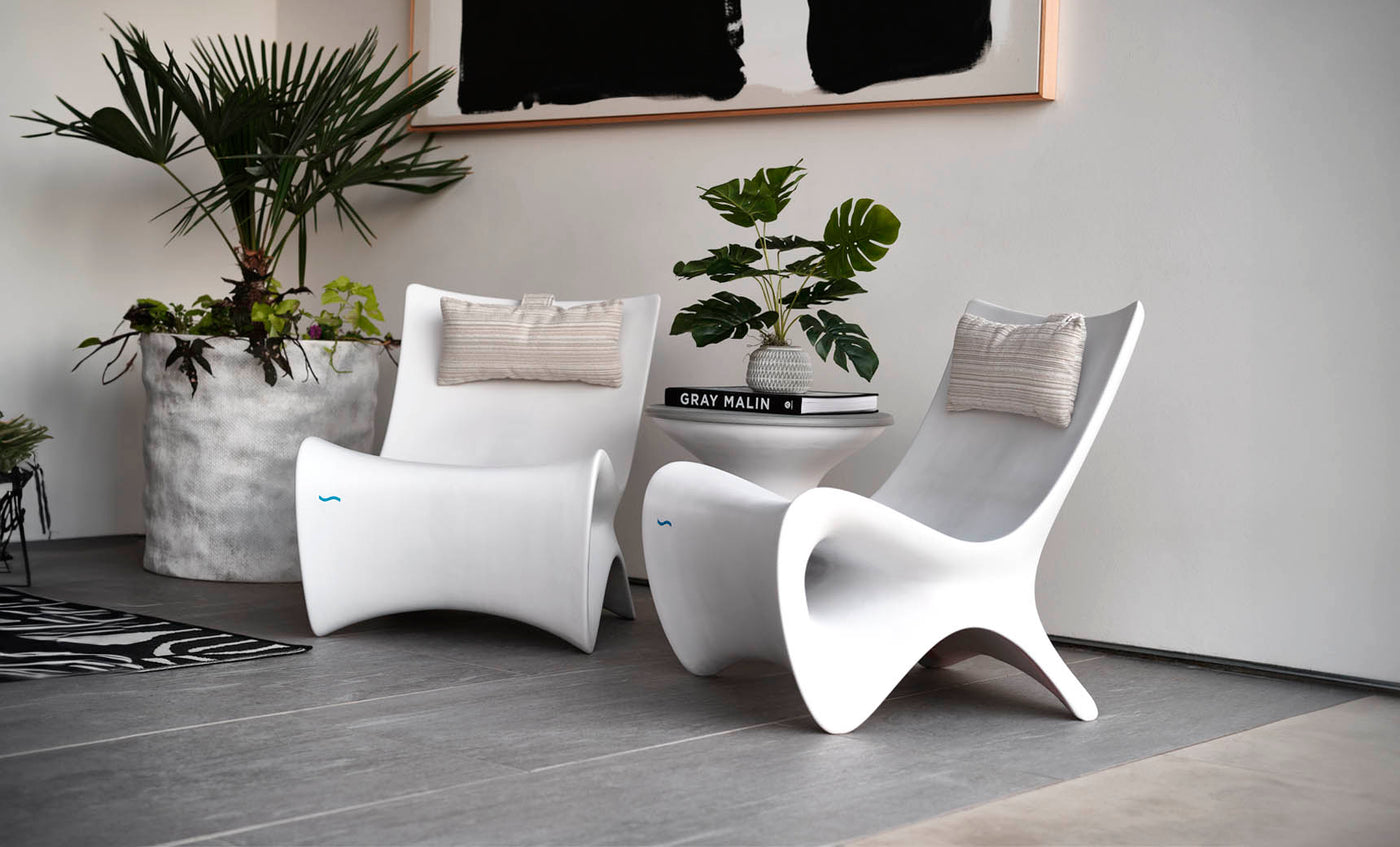 A modern interior features two sleek white chairs with beige cushions, centered around a small white table holding a stack of books and a potted plant. Large potted greenery sits nearby, and an abstract black-and-white artwork hangs above.