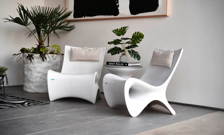 A modern interior features two sleek white chairs with beige cushions, centered around a small white table holding a stack of books and a potted plant. Large potted greenery sits nearby, and an abstract black-and-white artwork hangs above.