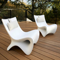 Two Autograph Chairs by Ledge Lounger, in modern white and complete with pillows, enhance a wooden deck surrounded by metal railings, providing breathtaking views of lush green trees for peaceful outdoor relaxation.