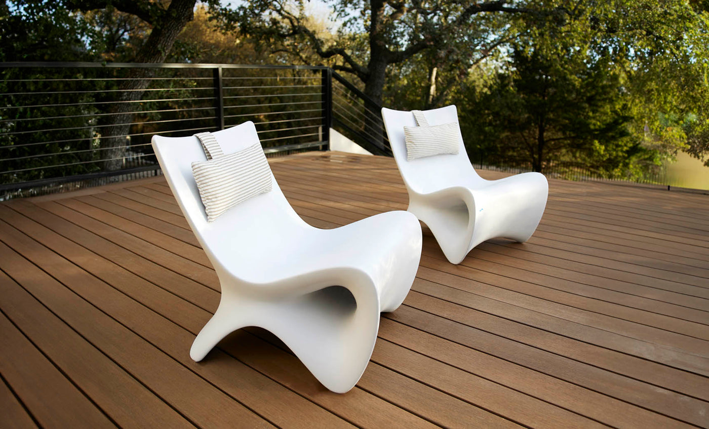 Two Autograph Chairs by Ledge Lounger, in modern white and complete with pillows, enhance a wooden deck surrounded by metal railings, providing breathtaking views of lush green trees for peaceful outdoor relaxation.