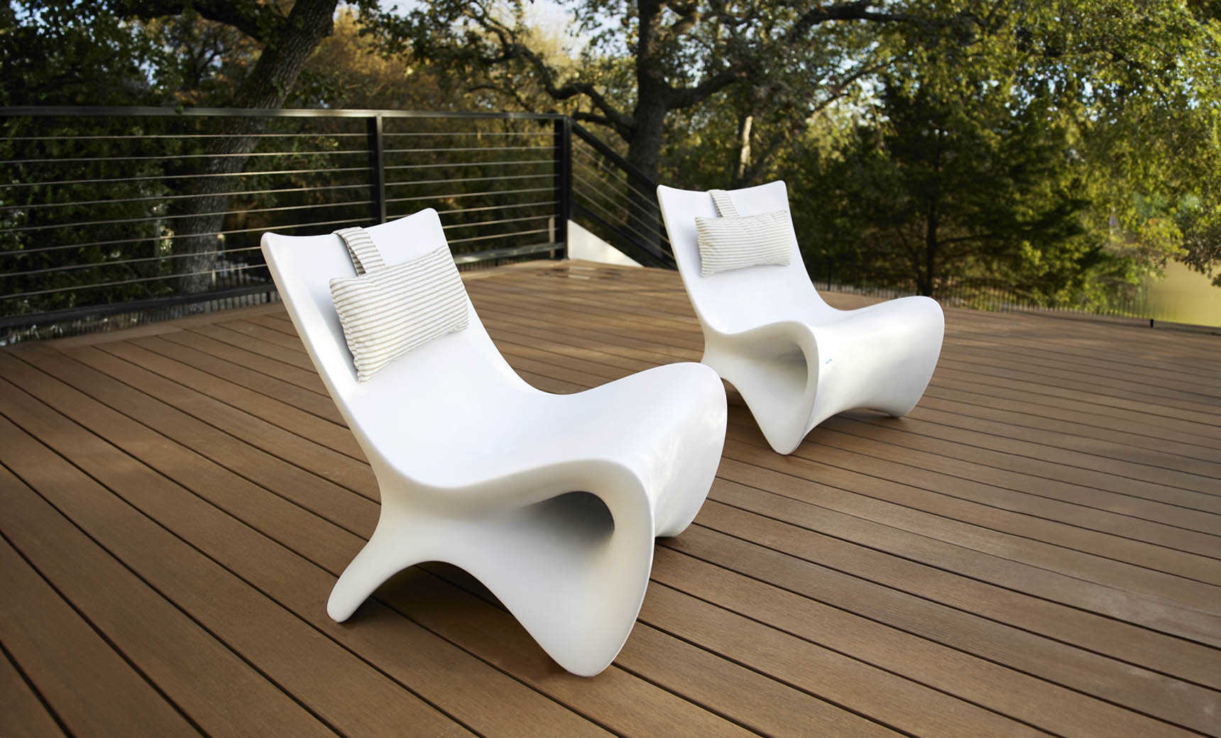 Two Autograph Chairs by Ledge Lounger, in modern white and complete with pillows, enhance a wooden deck surrounded by metal railings, providing breathtaking views of lush green trees for peaceful outdoor relaxation.