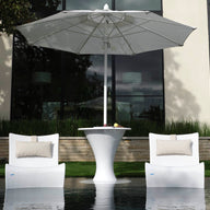 Two Ledge Lounger Autograph Chairs with beige pillows face a sleek white table under a large umbrella outdoors, set by reflective water and enhanced by a contemporary building window in the serene backdrop.