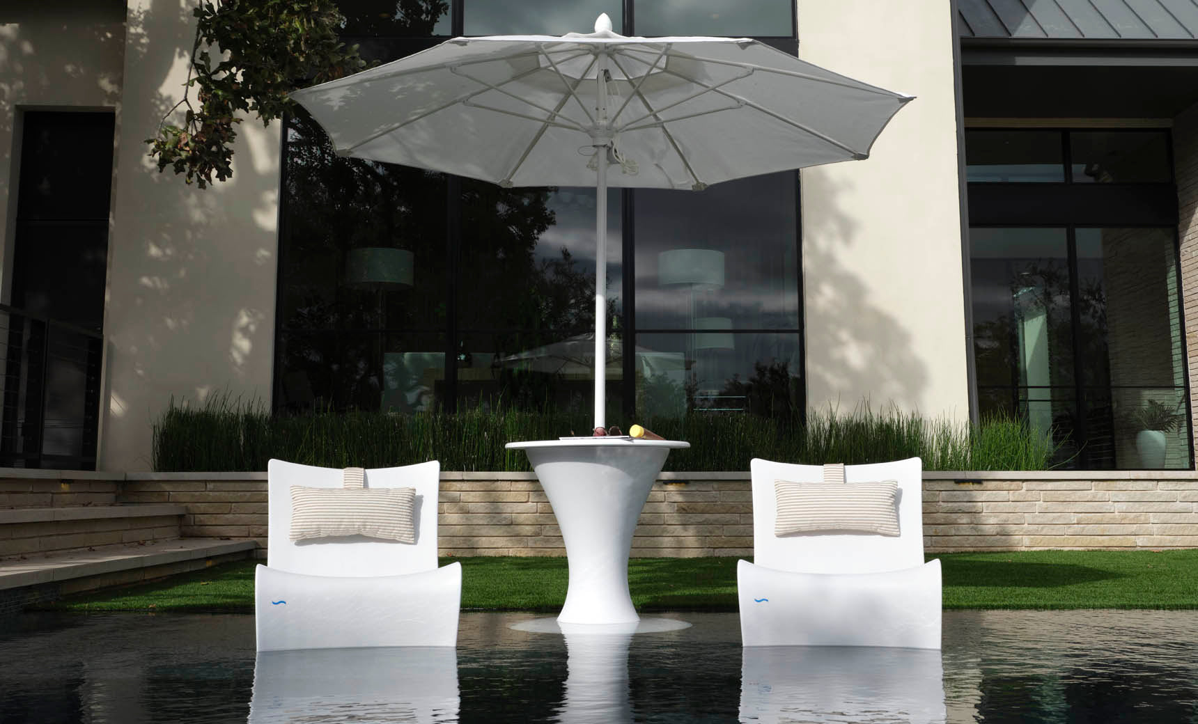 Two Ledge Lounger Autograph Chairs with beige pillows face a sleek white table under a large umbrella outdoors, set by reflective water and enhanced by a contemporary building window in the serene backdrop.