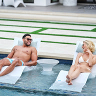 A man and woman enjoy a luxurious lounging experience on partially submerged Ledge Lounger Autograph Chaises. Clad in swimsuits and sunglasses, they smile over a small round table, with a modern patio enhancing the serene outdoor backdrop.