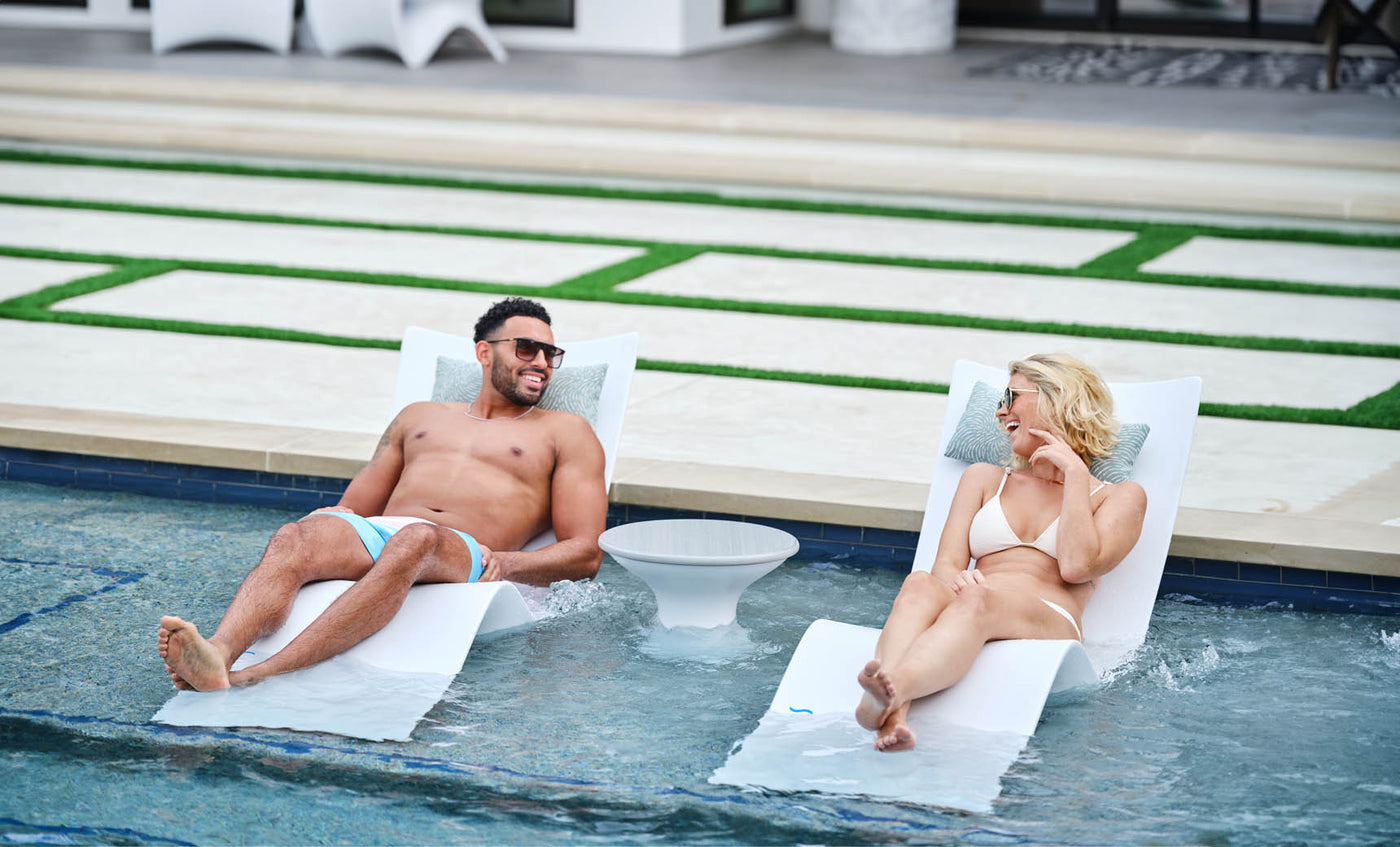 A man and woman enjoy a luxurious lounging experience on partially submerged Ledge Lounger Autograph Chaises. Clad in swimsuits and sunglasses, they smile over a small round table, with a modern patio enhancing the serene outdoor backdrop.