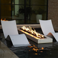 Two Ledge Lounger Autograph Chaises rest in a shallow pool beside a rectangular fire pit with flames. Positioned near a brick wall and large window, they create a modern and relaxing outdoor lounging area.