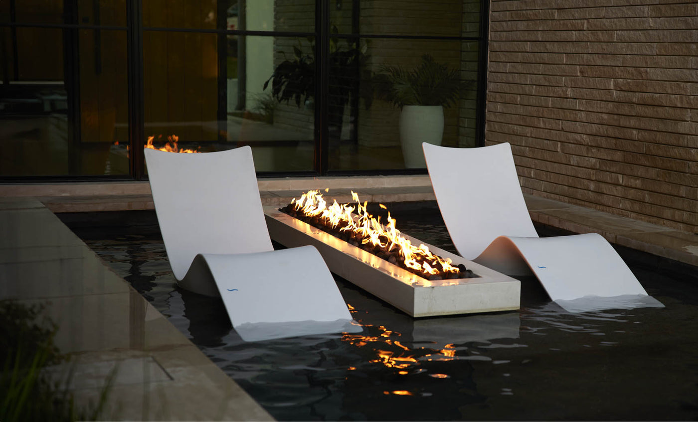 Two Ledge Lounger Autograph Chaises rest in a shallow pool beside a rectangular fire pit with flames. Positioned near a brick wall and large window, they create a modern and relaxing outdoor lounging area.