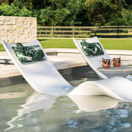 Two white Ledge Lounger Autograph Chaise lounges with green leaf-patterned cushions are partially submerged in a pool. A small table between them holds drinks and snacks, set against a backdrop of a stone wall, lush trees, and wooden fence for an idyllic lounging experience.