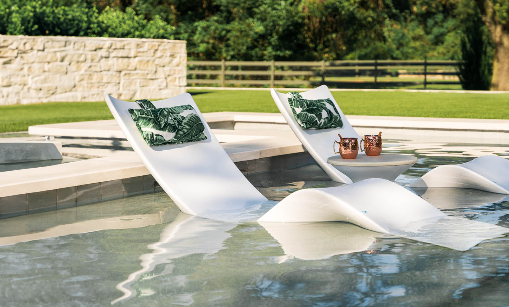 Two white Ledge Lounger Autograph Chaise lounges with green leaf-patterned cushions are partially submerged in a pool. A small table between them holds drinks and snacks, set against a backdrop of a stone wall, lush trees, and wooden fence for an idyllic lounging experience.