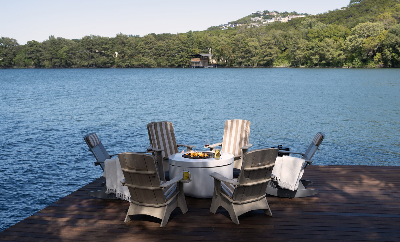 Six Ledge Lounger Adirondack chairs surround a fire pit with a glass top on a deck by a calm lake. Hillside homes and trees enhance the view. Mainstay Adirondack Cushions with white blankets add coziness.
