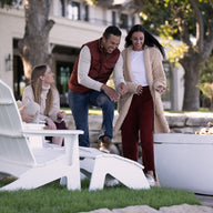 Four people enjoy outdoor relaxation by a fire pit. Two sit on Ledge Loungers white Legacy Adirondack Ottomans while the others stand nearby, engrossed in conversation. Trees and a building backdrop enhance the cozy atmosphere, creating a friendly and relaxed mood.