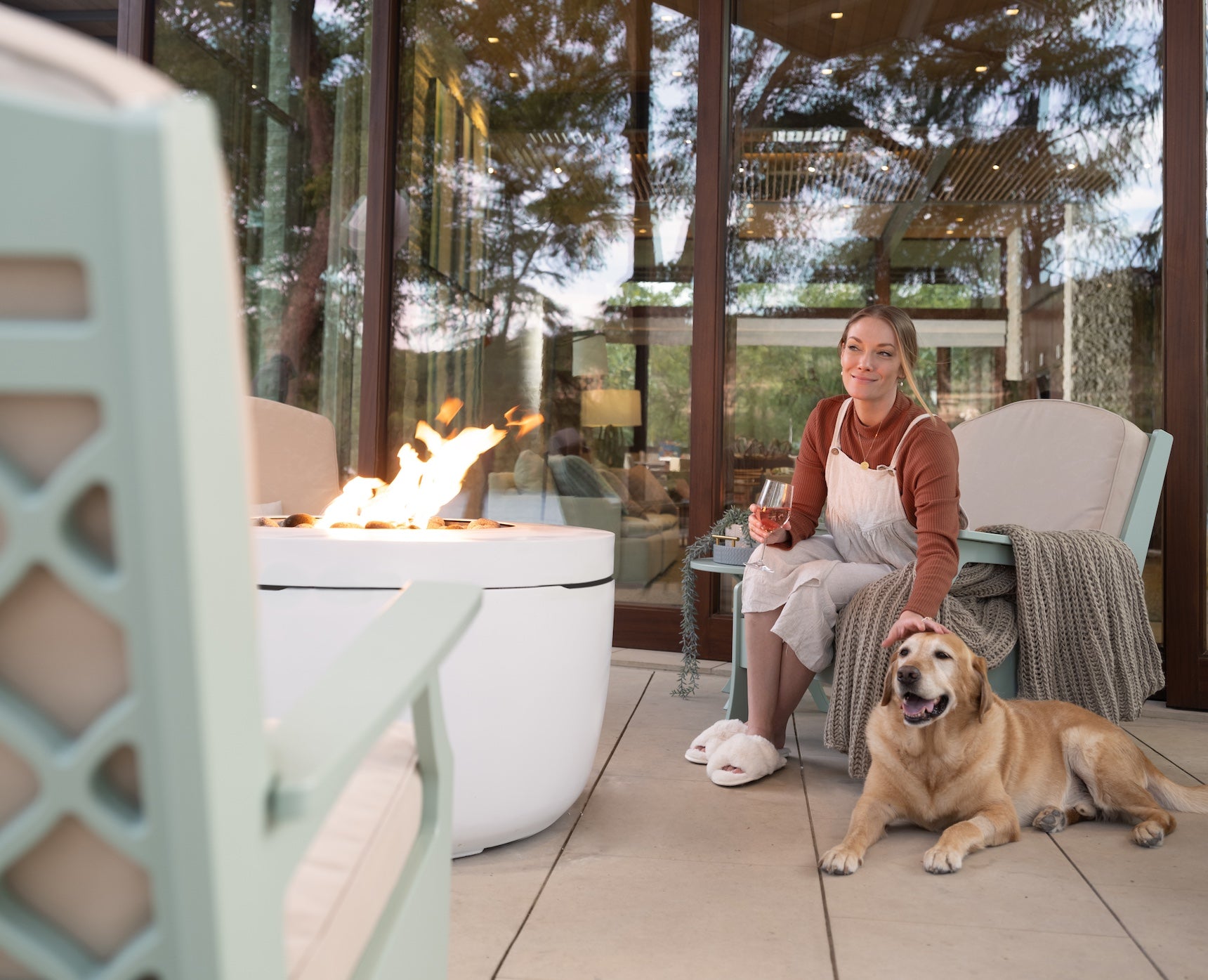 A woman sits on a patio chair, holding a glass of wine and petting a dog. Theres a fire pit next to her with flames visible. She is relaxed, smiling, and wearing comfortable clothing. The setting appears cozy and inviting, with trees reflected in glass doors.