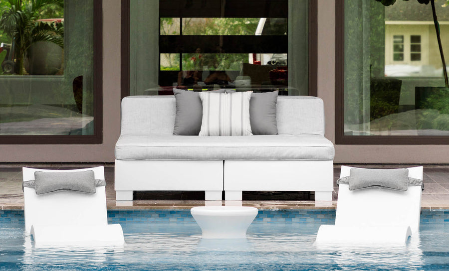 Two Affinity Square Sunbeds with backrest cushions from Ledge Lounger sit partially submerged poolside, facing a modern sofa with gray and white cushions. A small round table nestles between them, while large glass doors reflect the lush greenery beyond.