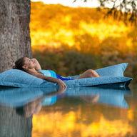 A person with long hair relaxes on a Ledge Lounger Laze Pillow in a serene body of water, surrounded by trees and a golden landscape under warm light, enjoying perfect outdoor comfort.