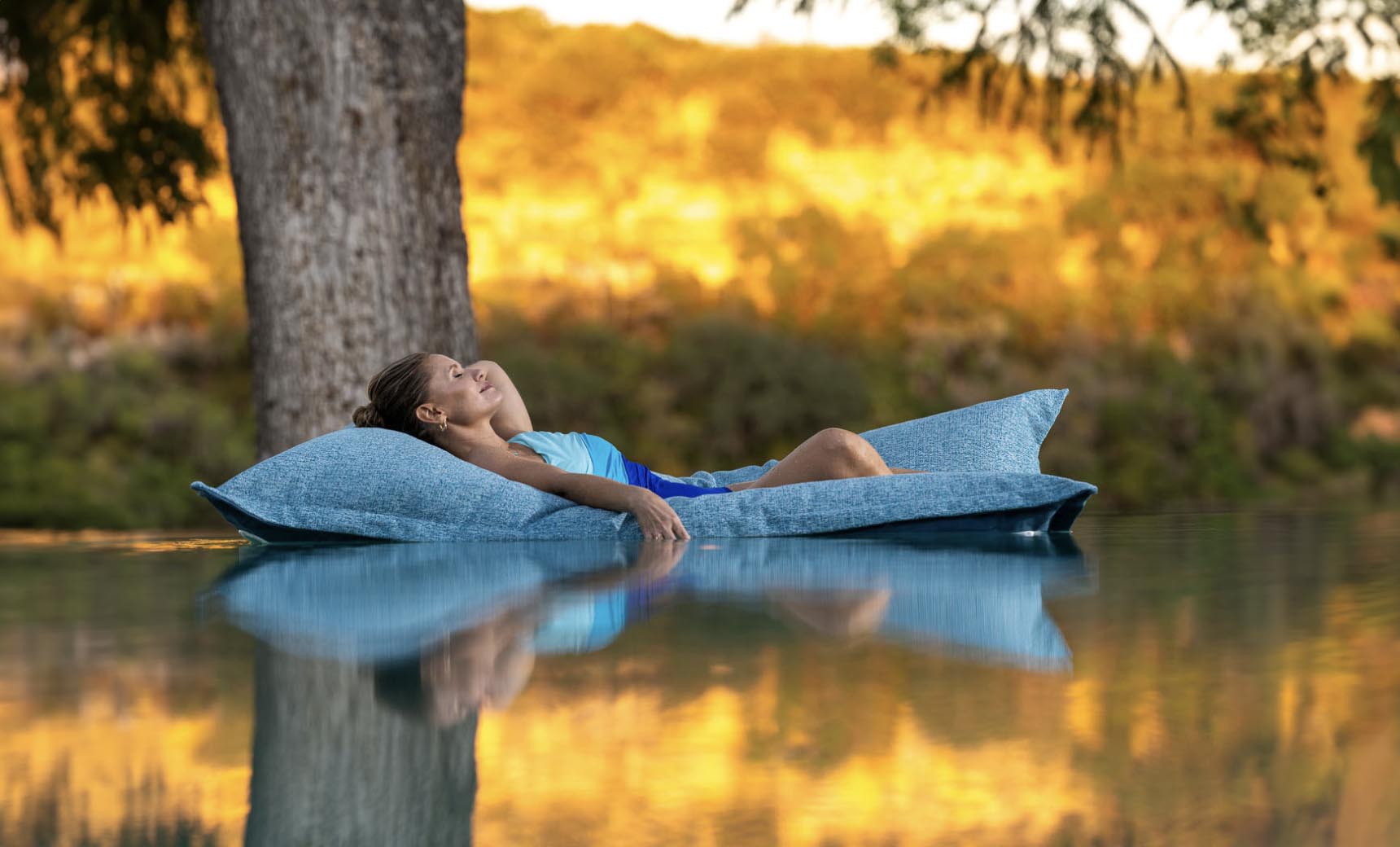 A person with long hair relaxes on a Ledge Lounger Laze Pillow in a serene body of water, surrounded by trees and a golden landscape under warm light, enjoying perfect outdoor comfort.