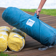 A barefoot individual sets a blue, cylindrical Laze Hammock by Ledge Lounger next to two yellow-patterned rolls on a wooden deck near a grassy spot, with water shimmering in the background, suggesting peaceful outdoor adventures.
