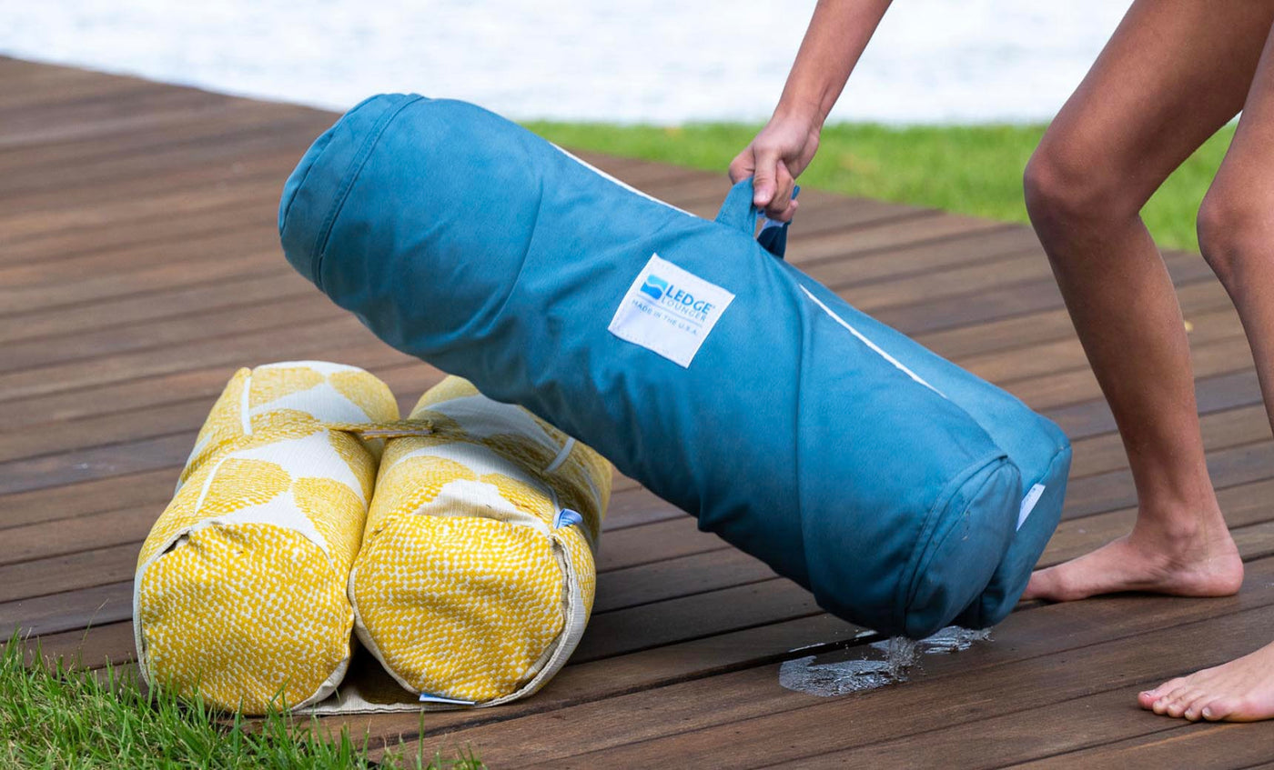 A barefoot individual sets a blue, cylindrical Laze Hammock by Ledge Lounger next to two yellow-patterned rolls on a wooden deck near a grassy spot, with water shimmering in the background, suggesting peaceful outdoor adventures.