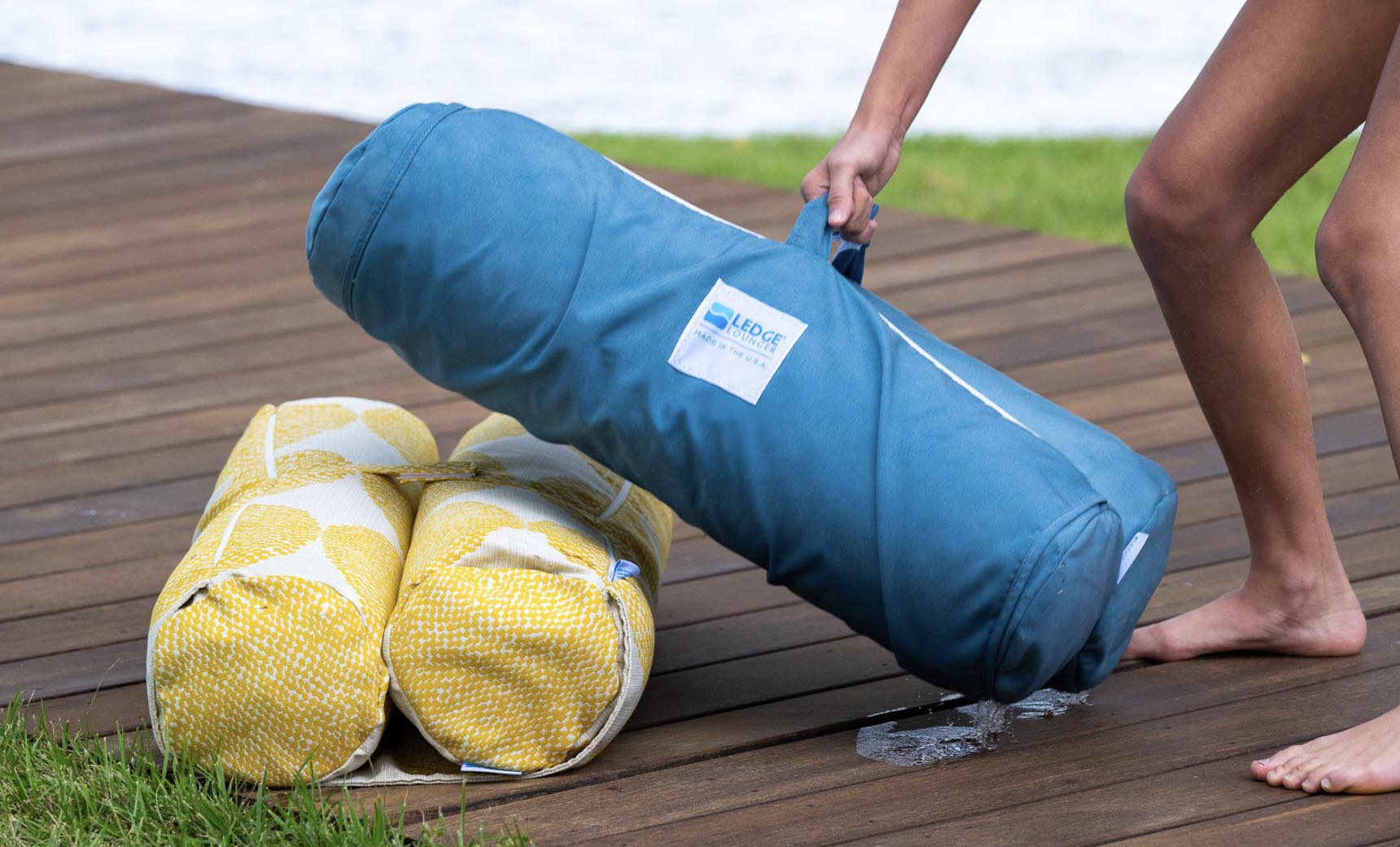 A barefoot individual sets a blue, cylindrical Laze Hammock by Ledge Lounger next to two yellow-patterned rolls on a wooden deck near a grassy spot, with water shimmering in the background, suggesting peaceful outdoor adventures.