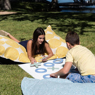 Two kids play a giant lawn game of Ledge Loungers Checkers & Tic Tac Toe on grass, relaxing on bean bags with circular yellow patterns. Trees and water form a serene backdrop under the clear sky.