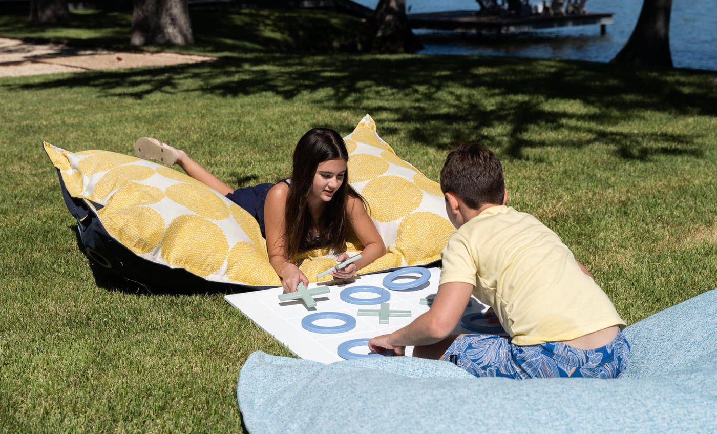 Two kids play a giant lawn game of Ledge Loungers Checkers & Tic Tac Toe on grass, relaxing on bean bags with circular yellow patterns. Trees and water form a serene backdrop under the clear sky.