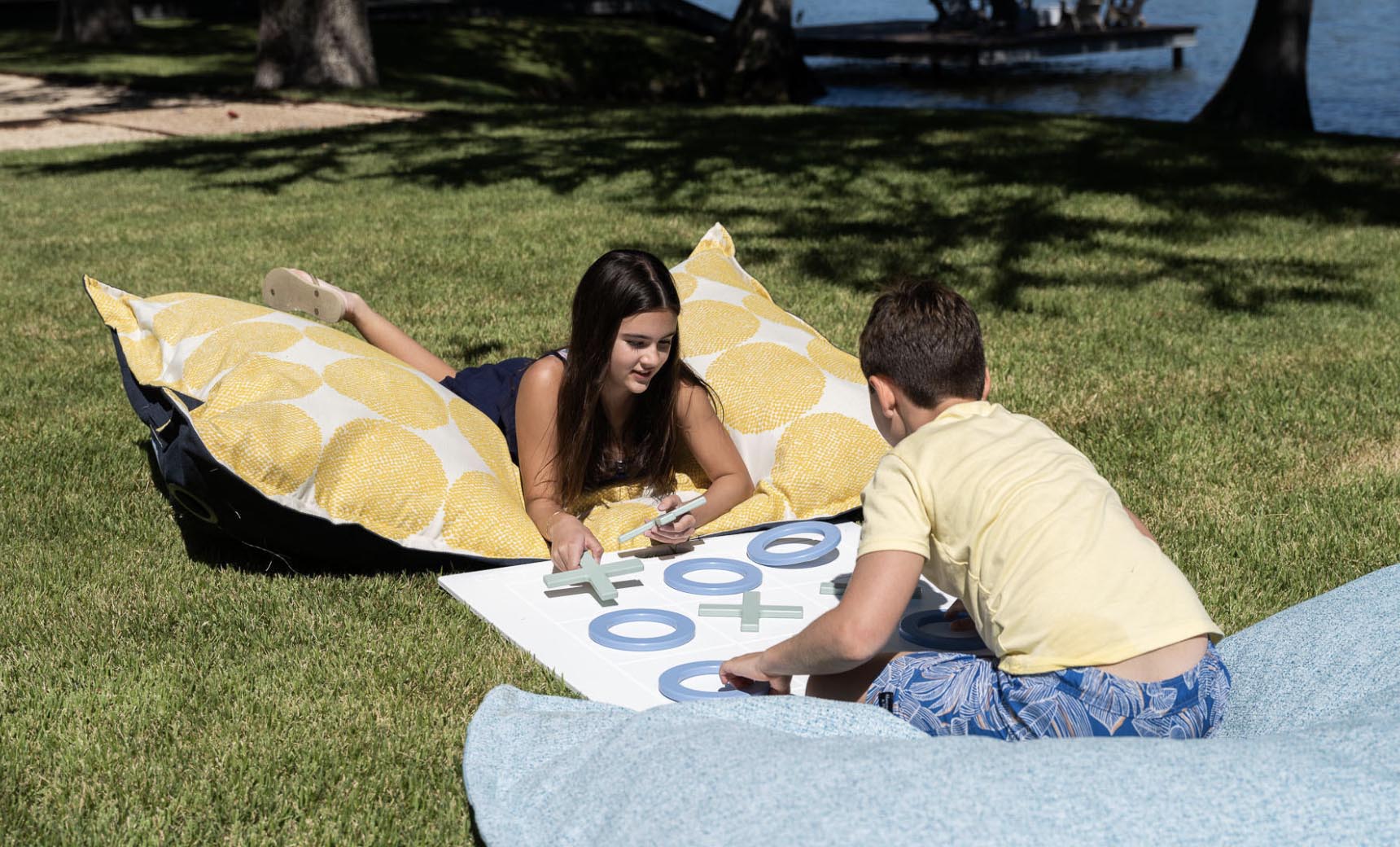 Two kids play a giant lawn game of Ledge Loungers Checkers & Tic Tac Toe on grass, relaxing on bean bags with circular yellow patterns. Trees and water form a serene backdrop under the clear sky.