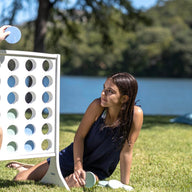 A boy and girl are enjoying oversized outdoor fun with Ledge Loungers Four In A Row by the lake. The boy inserts a disc on the giant board while the girl crouches nearby, observing him. Trees gently sway along the waters edge in the background.
