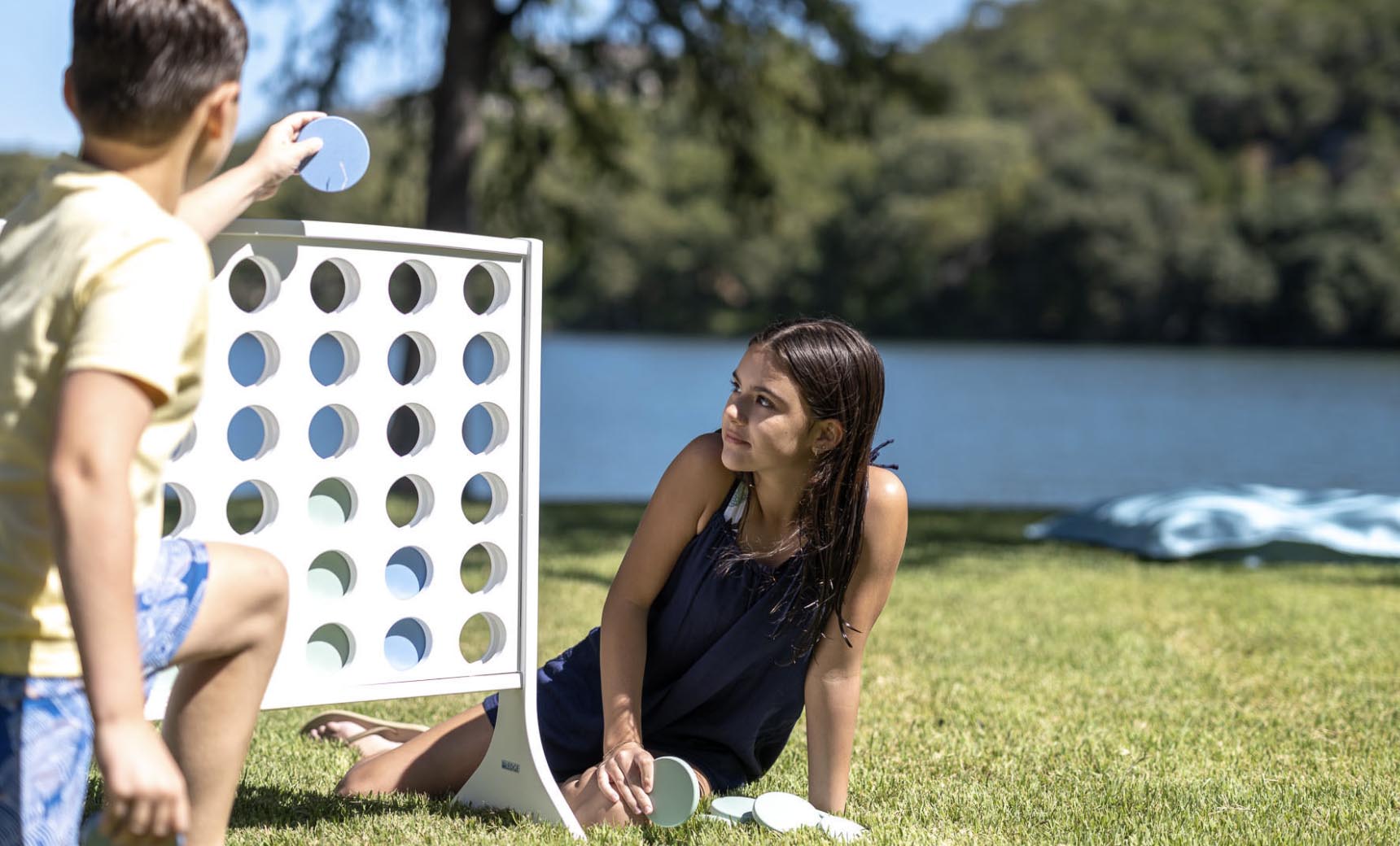 A boy and girl are enjoying oversized outdoor fun with Ledge Loungers Four In A Row by the lake. The boy inserts a disc on the giant board while the girl crouches nearby, observing him. Trees gently sway along the waters edge in the background.
