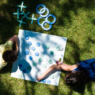 Two children in a vibrant scene play an immersive game of Checkers & Tic Tac Toe by Ledge Lounger on the grass. One wears a yellow shirt, the other a blue dress. The board displays scattered pieces, extra circles, and crosses, with tree-filtered sunlight enhancing the playful atmosphere.