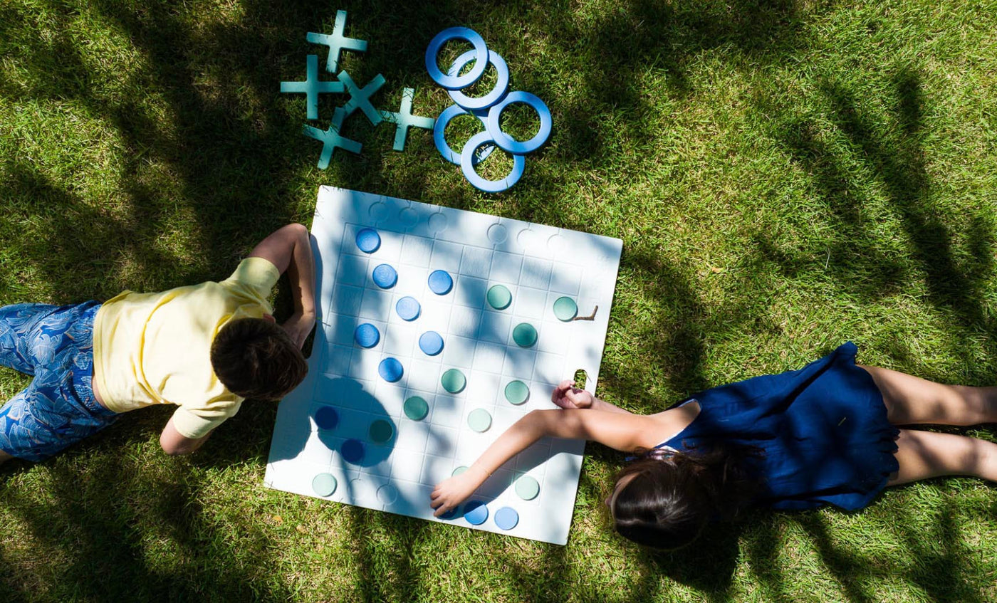 Two children in a vibrant scene play an immersive game of Checkers & Tic Tac Toe by Ledge Lounger on the grass. One wears a yellow shirt, the other a blue dress. The board displays scattered pieces, extra circles, and crosses, with tree-filtered sunlight enhancing the playful atmosphere.